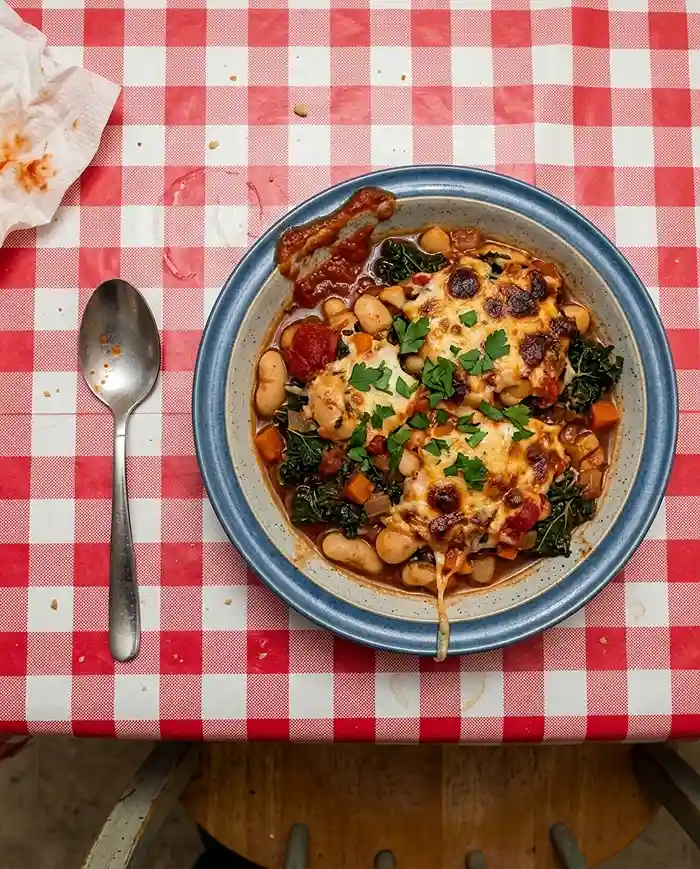 beans, tomato, kale under a brown bubbly cheese crust broken into uneven patches, parsley scatter, cheese string down the outside, dome light
