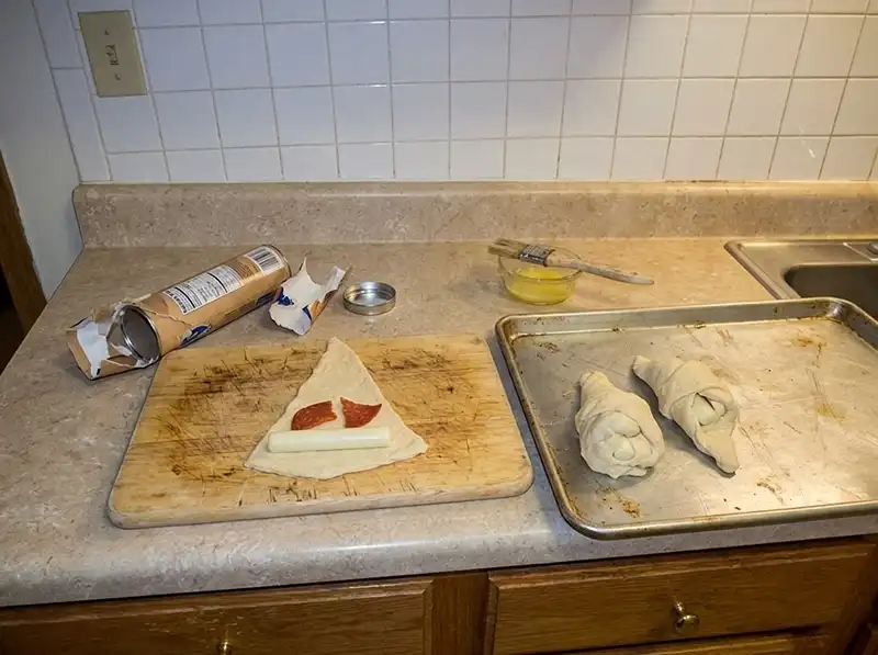 cheese stick laid across the wide end of a triangle with pepperoni on top, two cones already rolled on the sheet, seams only partly sealed, one splitting with cheese peeking out