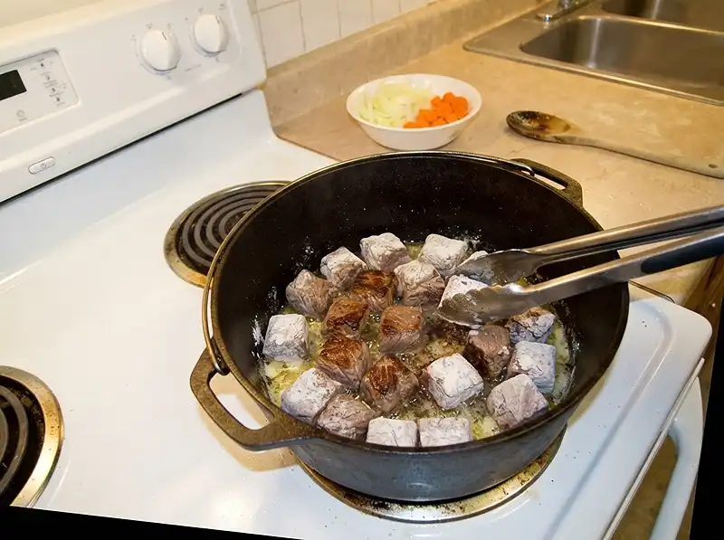 Guinness Pie with Tender Beef 4 flour-coated beef browning in the cast-iron Dutch oven, some faces deep brown and others still pale grey, butter and oil shimmering, onion and carrot waiting in a bowl