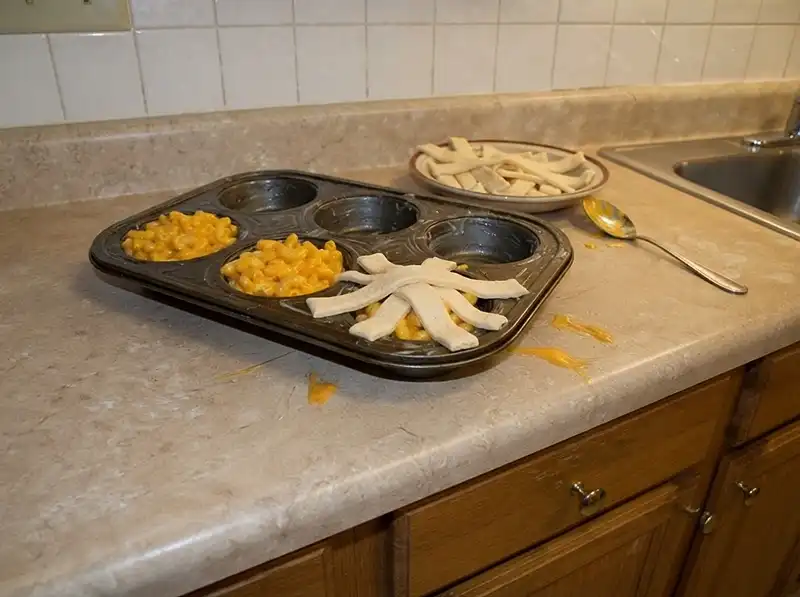 greased muffin tin, three cups filled with saucy mac, raw flabby dough strips being laid criss-cross on one, most cups still bare, no eyes yet