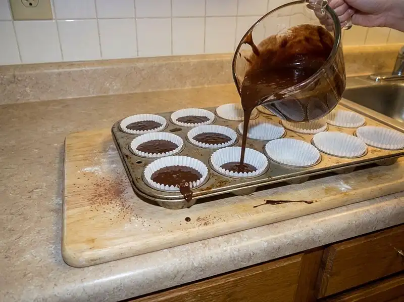 thin dark batter poured from a measuring cup into paper liners in the muffin tin, only half full, uneven, one dripped down the tin